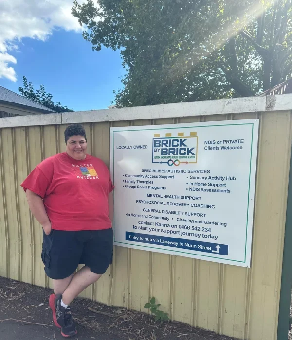 Happy support worker leaning against fence next to Brick by Brick sign, smiling at camera