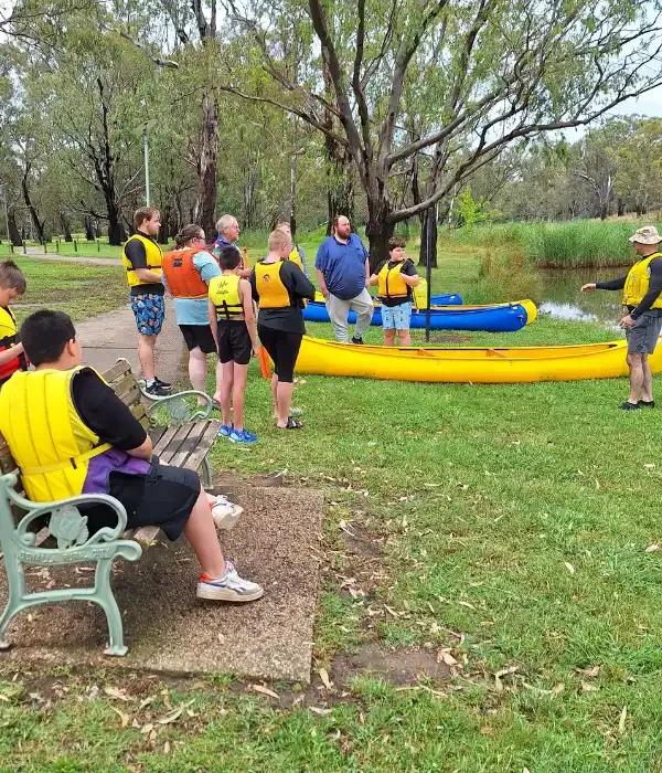 Group of people standing on the river bank with canoes, listening to instructor